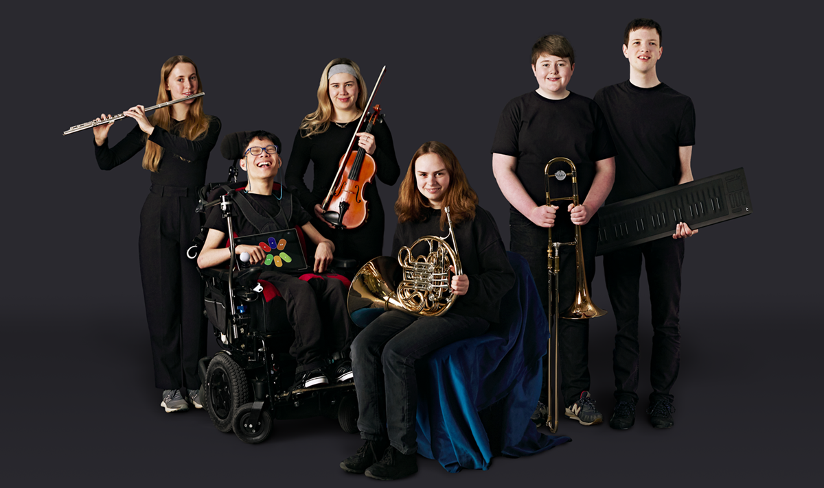 Six disabled and non-disabled musicians are holding their brass instruments. One who is a wheelchair user is showing an iPad screen with colourful shapes on it, which is his Clarion accessible instrument. Another musician is holding an electronic keyboard.