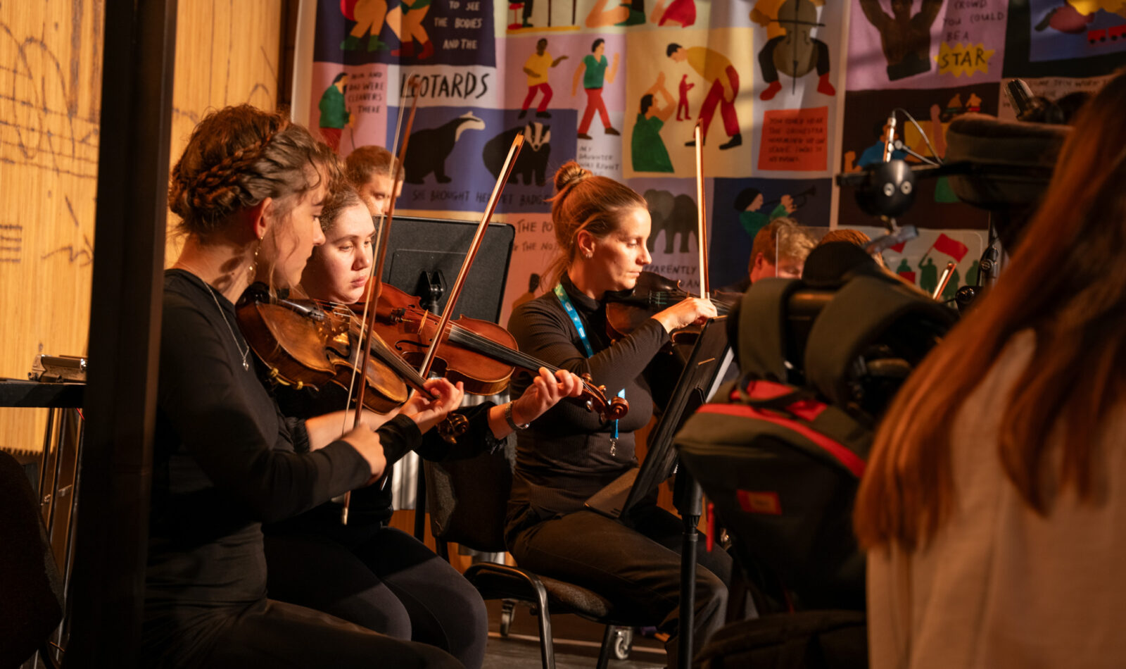 Three female violinists play on stage. There are other musicians on stage with them.