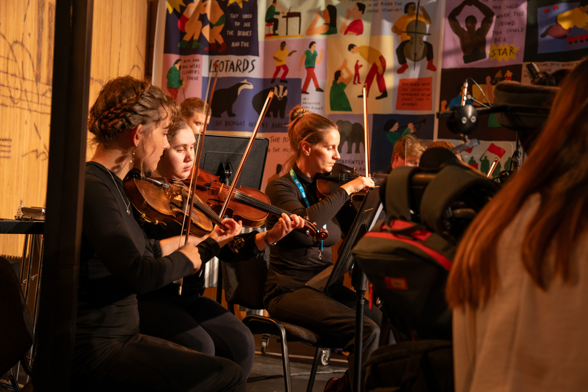 Three female violinists play on stage. There are other musicians on stage with them.