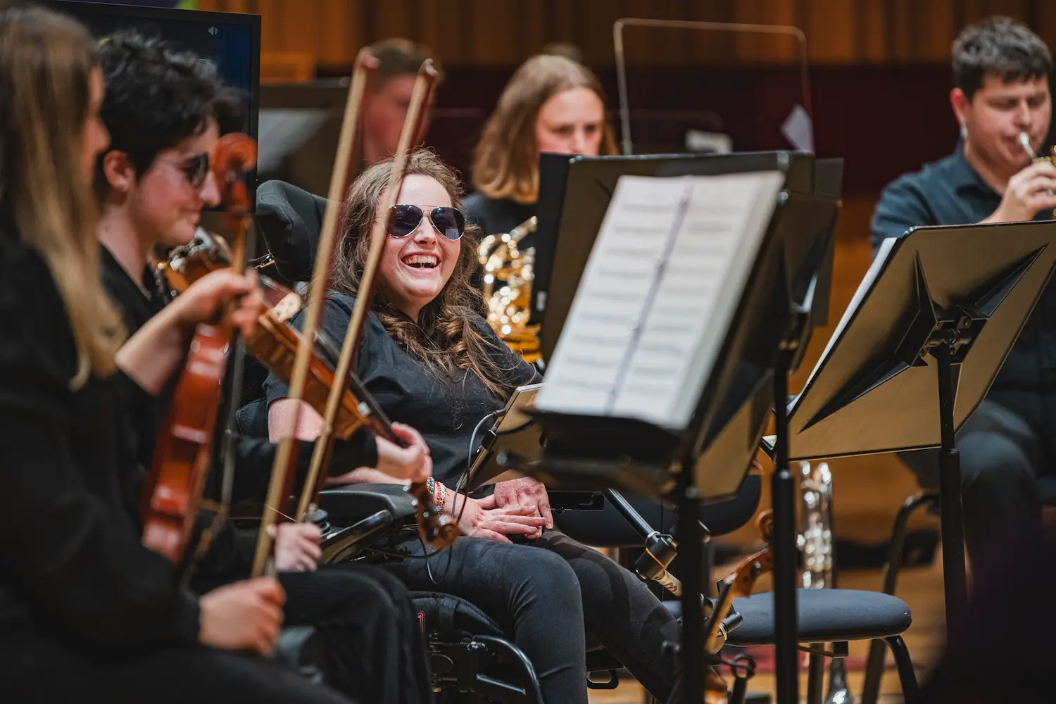 A young musician using a wheelchair smiles at an audience. She wears sunglasses and is surrounded by other NOYO musicians.