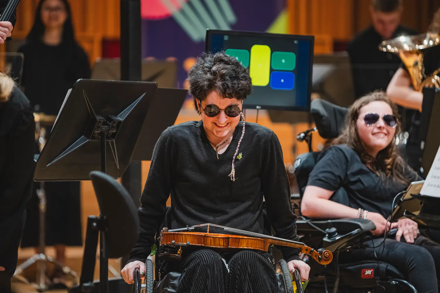 A young musician wearing sunglasses and using a wheelchair laughs onstage with NOYO as an audience applauds.