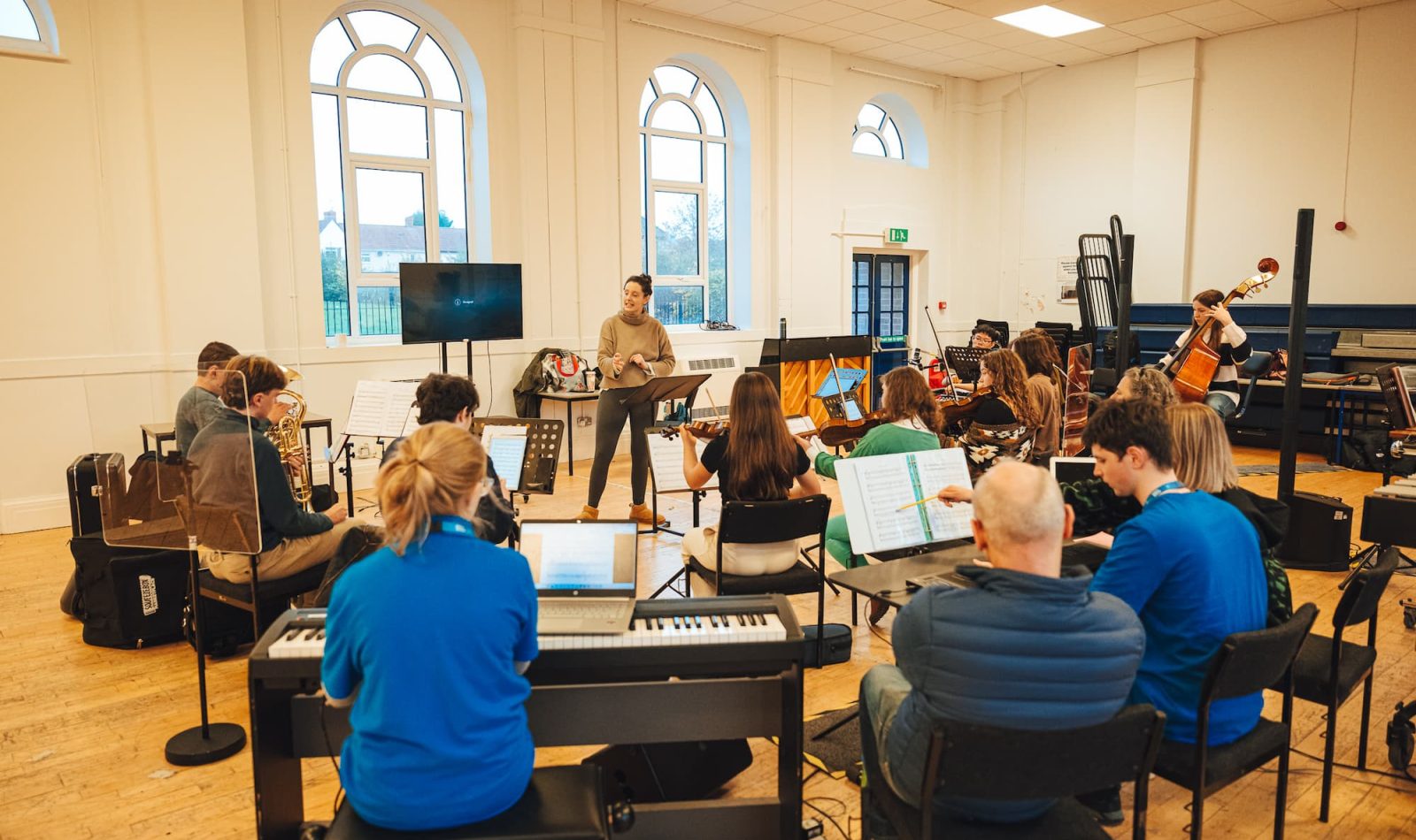 The Bristol NOYO Ensemble rehearse in a bright room with big windows. The ensemble is made up of disabled and non-disabled musicians playing digital, acoustic and accessible instruments.