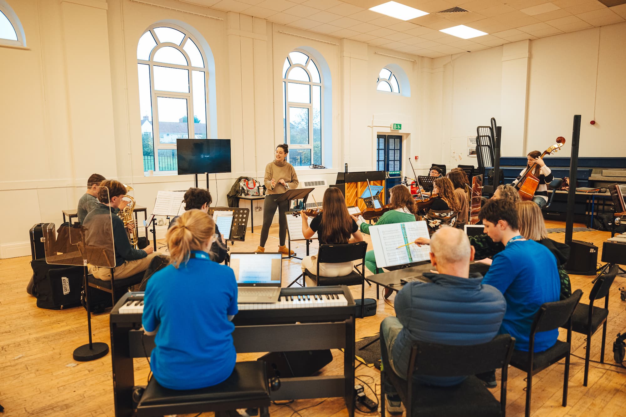 The Bristol NOYO Ensemble rehearse in a bright room with big windows. The ensemble is made up of disabled and non-disabled musicians playing digital, acoustic and accessible instruments.